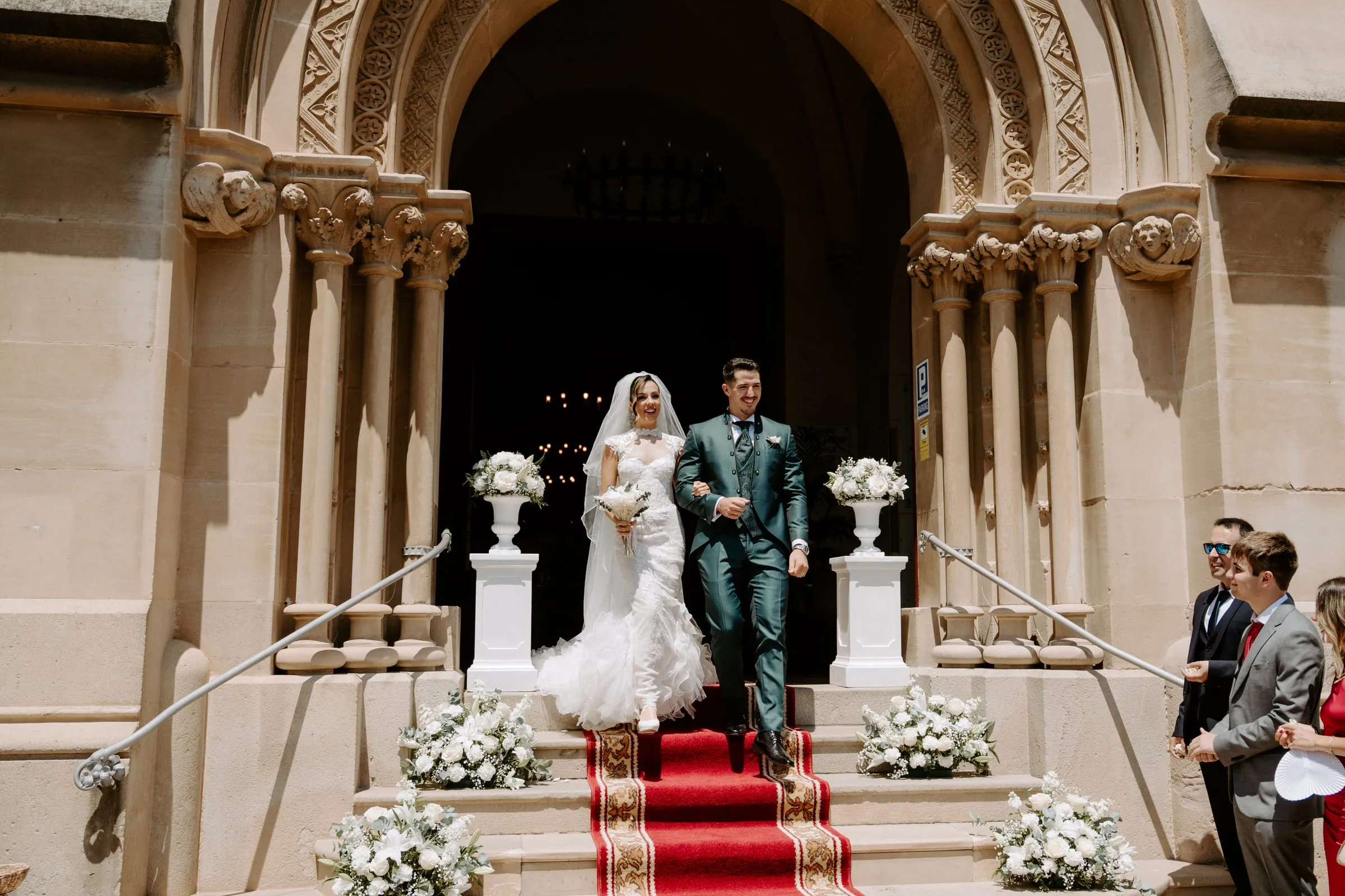 Los novios salen de la iglesia por la escalinata con alfombra roja, decorada con columnas y arreglos de flores blancas