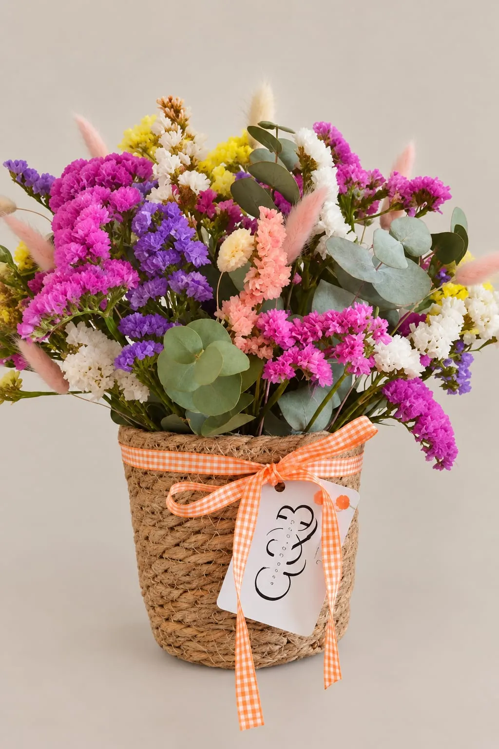 Cesta de fibra natural con flores preservadas de colores, statice morado y rosa, eucalipto y espigas, con un lazo de cuadros naranja y una tarjeta de Grow & Bloom.