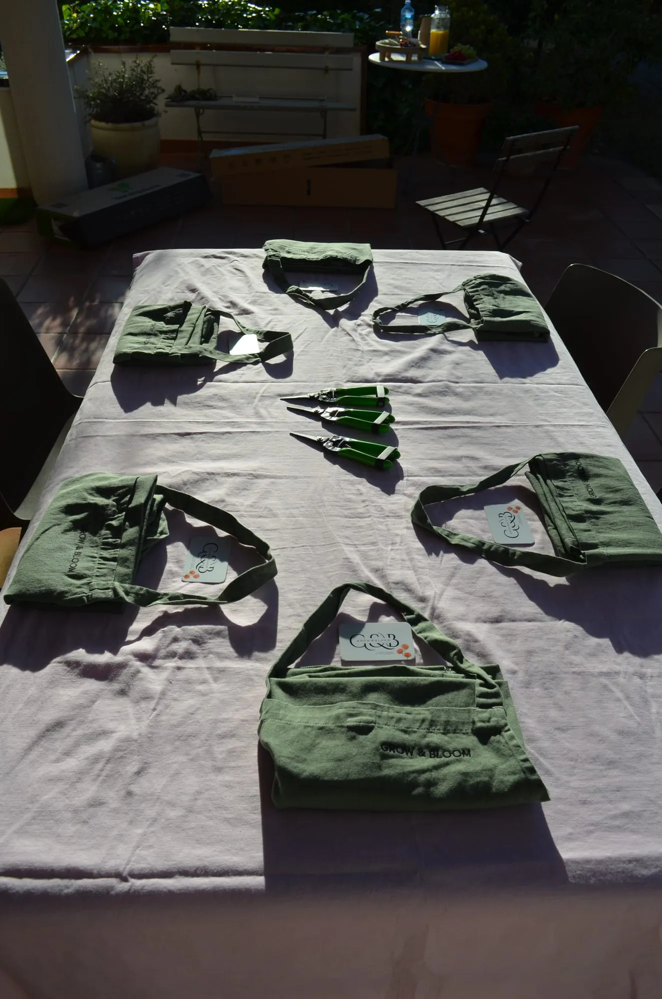 Mesa con mantel claro preparada para el taller, con delantales verdes de Grow & Bloom, tijeras de podar y tarjetas en cada sitio