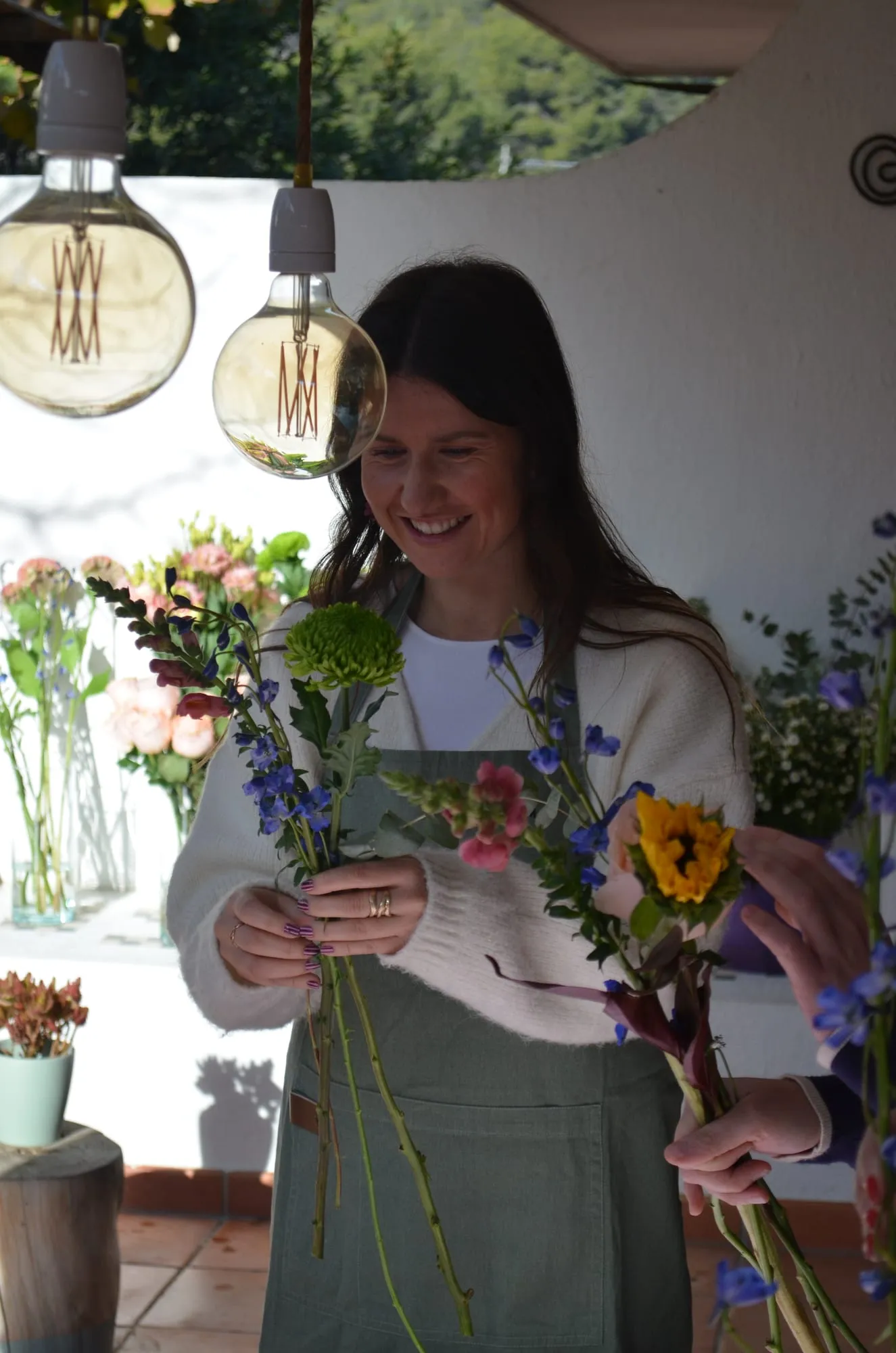 Una mujer sonríe mientras sostiene un ramo a medio montar con un crisantemo verde, espuela de caballero azul y un girasol