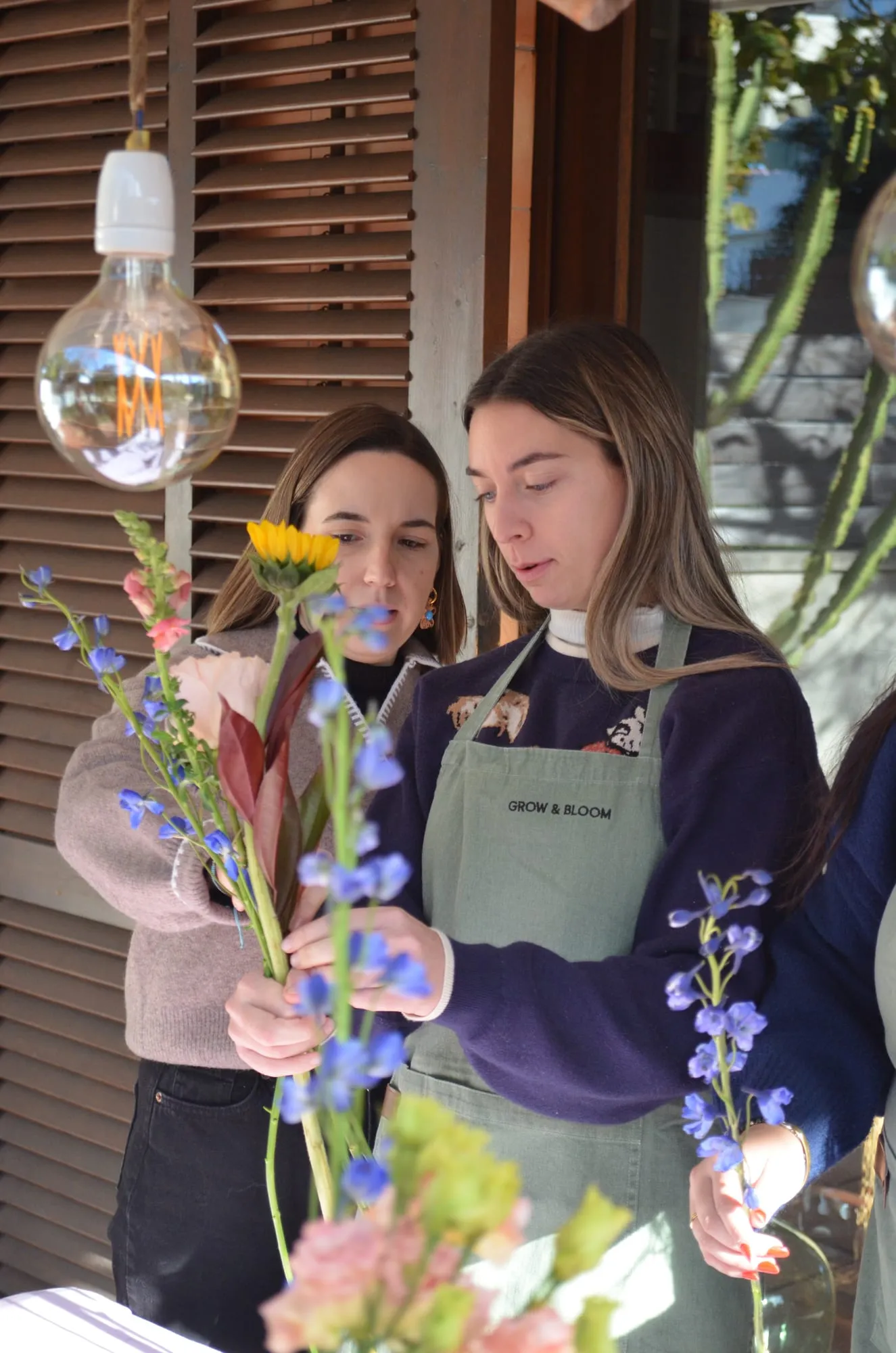 Dos chicas con delantal de Grow & Bloom montan sus ramos con espuela de caballero azul, un girasol y alstroemerias