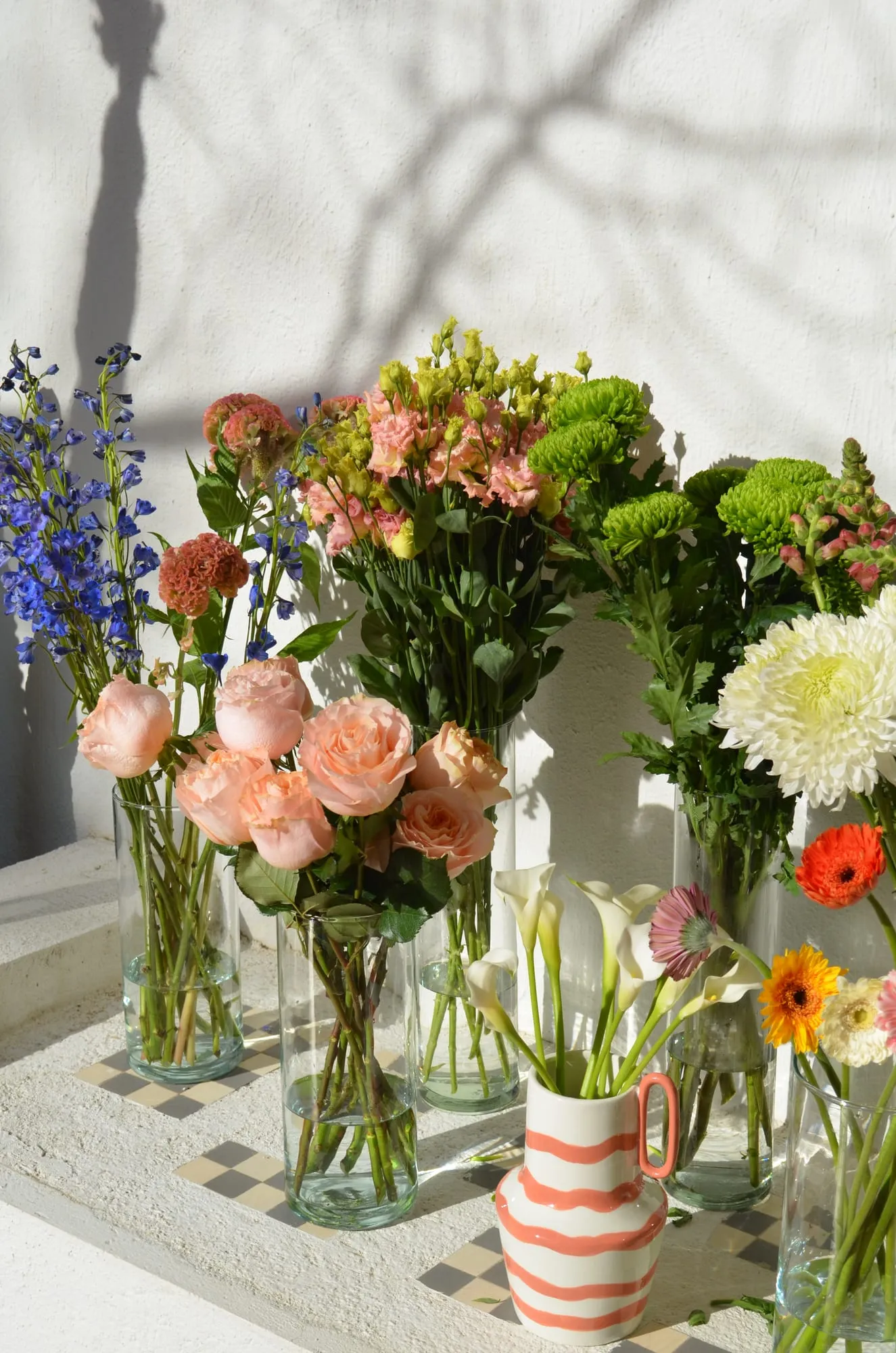 Jarrones de cristal y una jarra de rayas con rosas de jardín, crisantemos, calas, gerberas y espuela de caballero, al sol junto a una pared blanca
