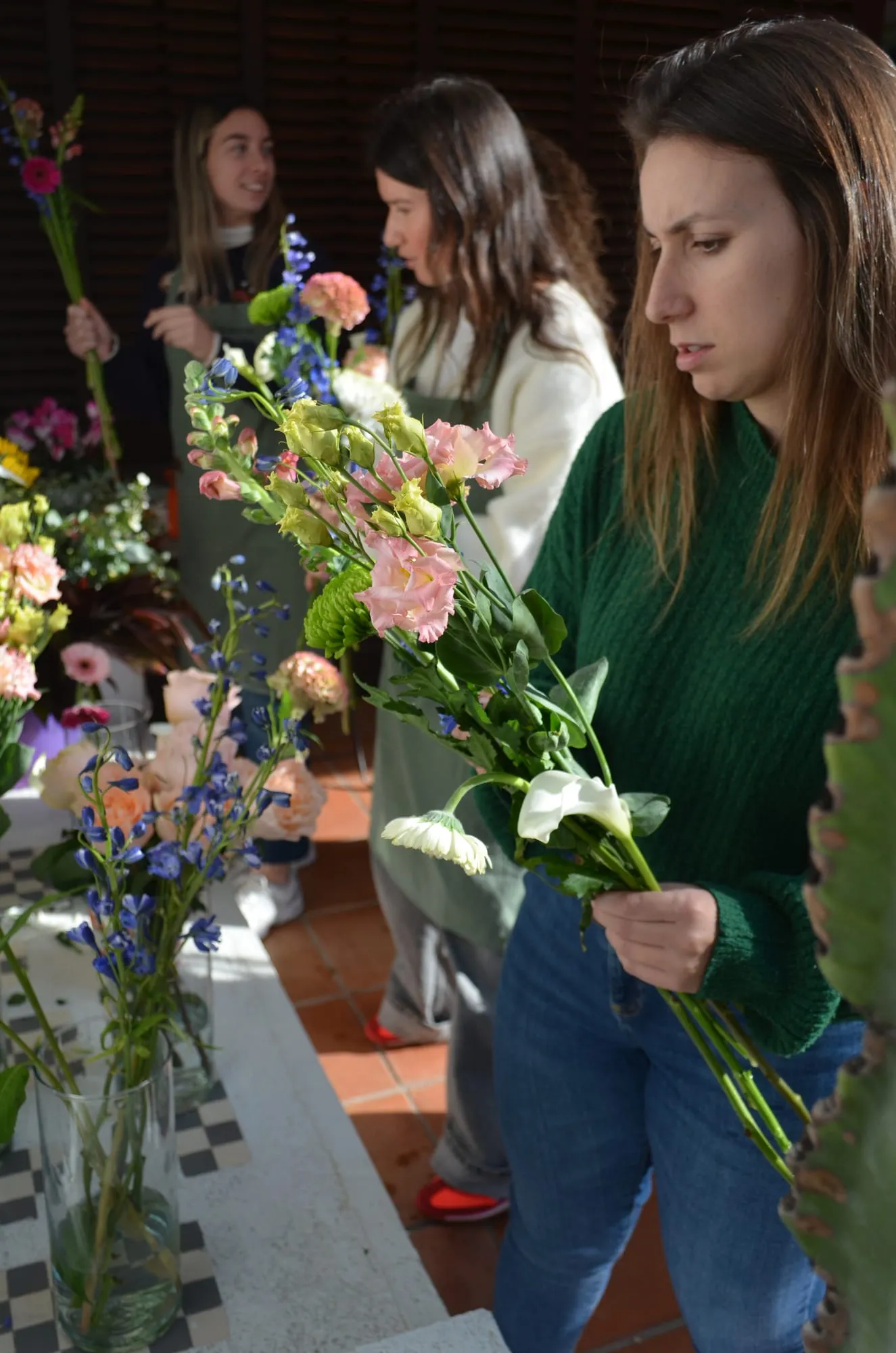 Varias chicas con delantal eligen flores de los jarrones; en primer plano, una de ellas con jersey verde sostiene su ramo