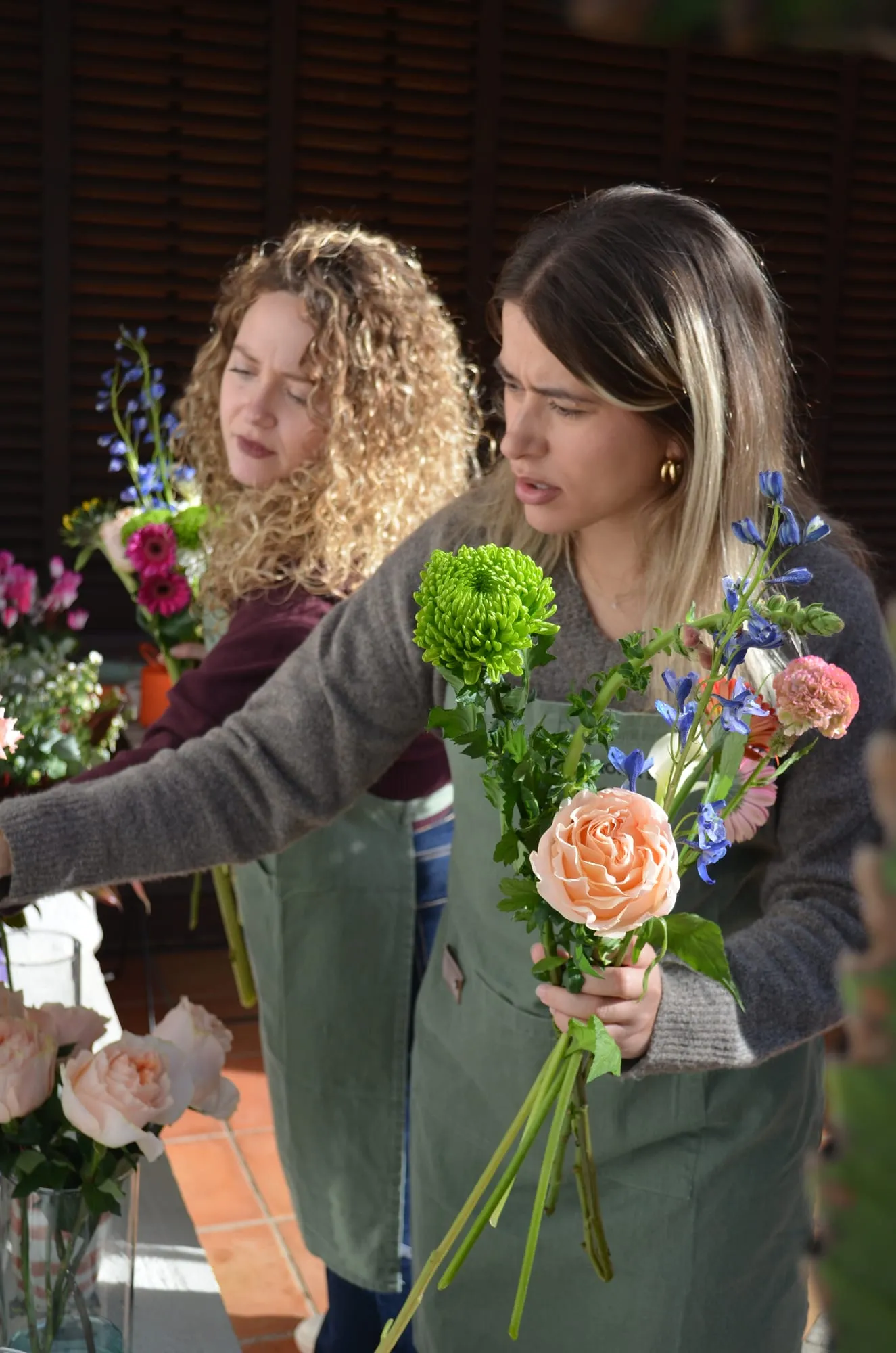 Una chica con delantal sostiene un ramo con una rosa de jardín melocotón y un crisantemo verde mientras otra coge flores detrás