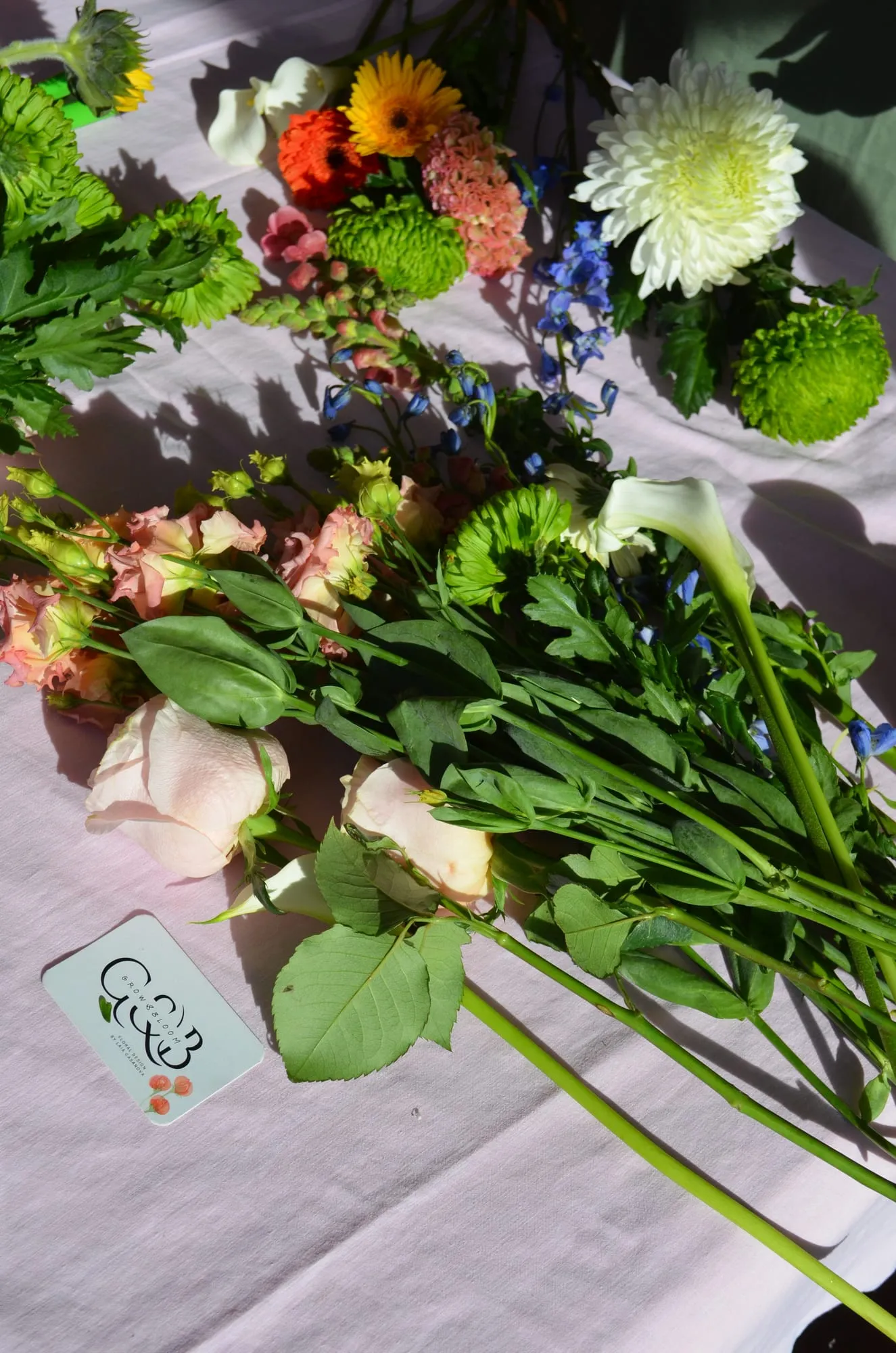 Flores cortadas sobre la mesa del taller: rosas, calas, una dalia blanca, gerberas y crisantemos verdes, junto a una tarjeta de Grow & Bloom