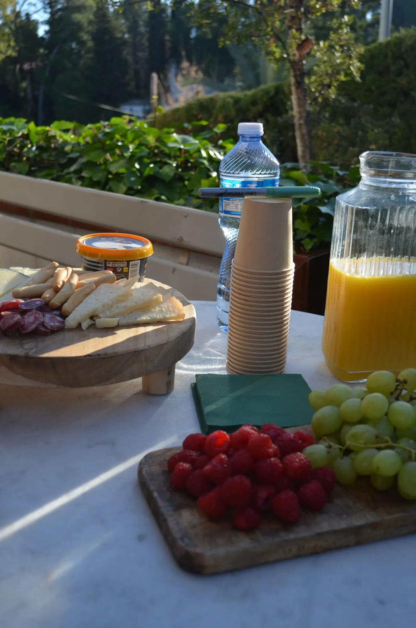 Pica pica al sol en la terraza: queso, embutido, frambuesas y uvas sobre tablas de madera, con zumo de naranja, agua y vasos de papel