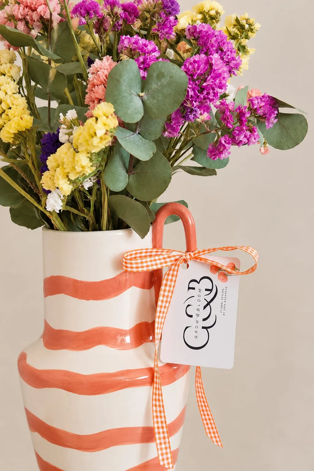 Flores preservadas de colores con eucalipto en un jarrón de cerámica blanco con rayas naranjas, con un lazo de cuadros y una tarjeta de Grow & Bloom.