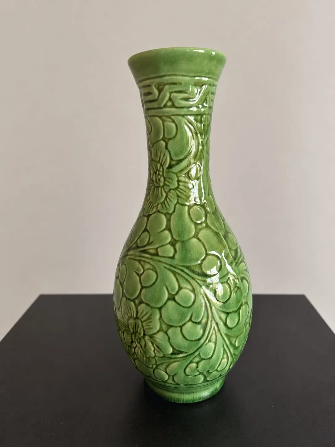 Jarrón de cerámica verde de cuello estrecho con flores en relieve y una greca en la parte superior.