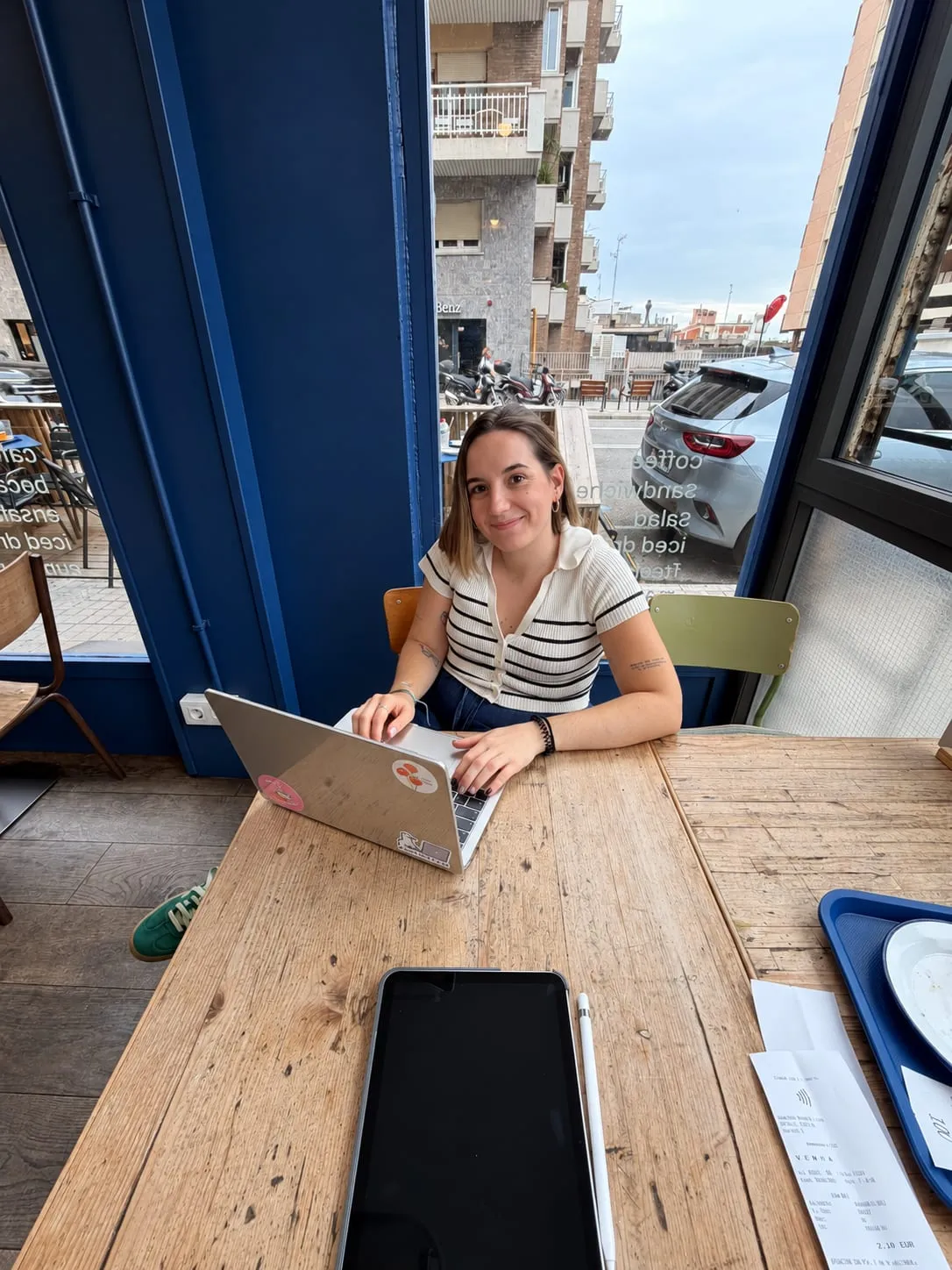 Laia sonríe sentada a una mesa de madera junto a una ventana, trabajando con su portátil.