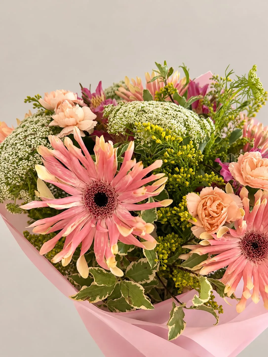 Detalle de un ramo de gerberas rosas, rosas color melocotón, solidago y flor de zanahoria, envuelto en papel rosa.