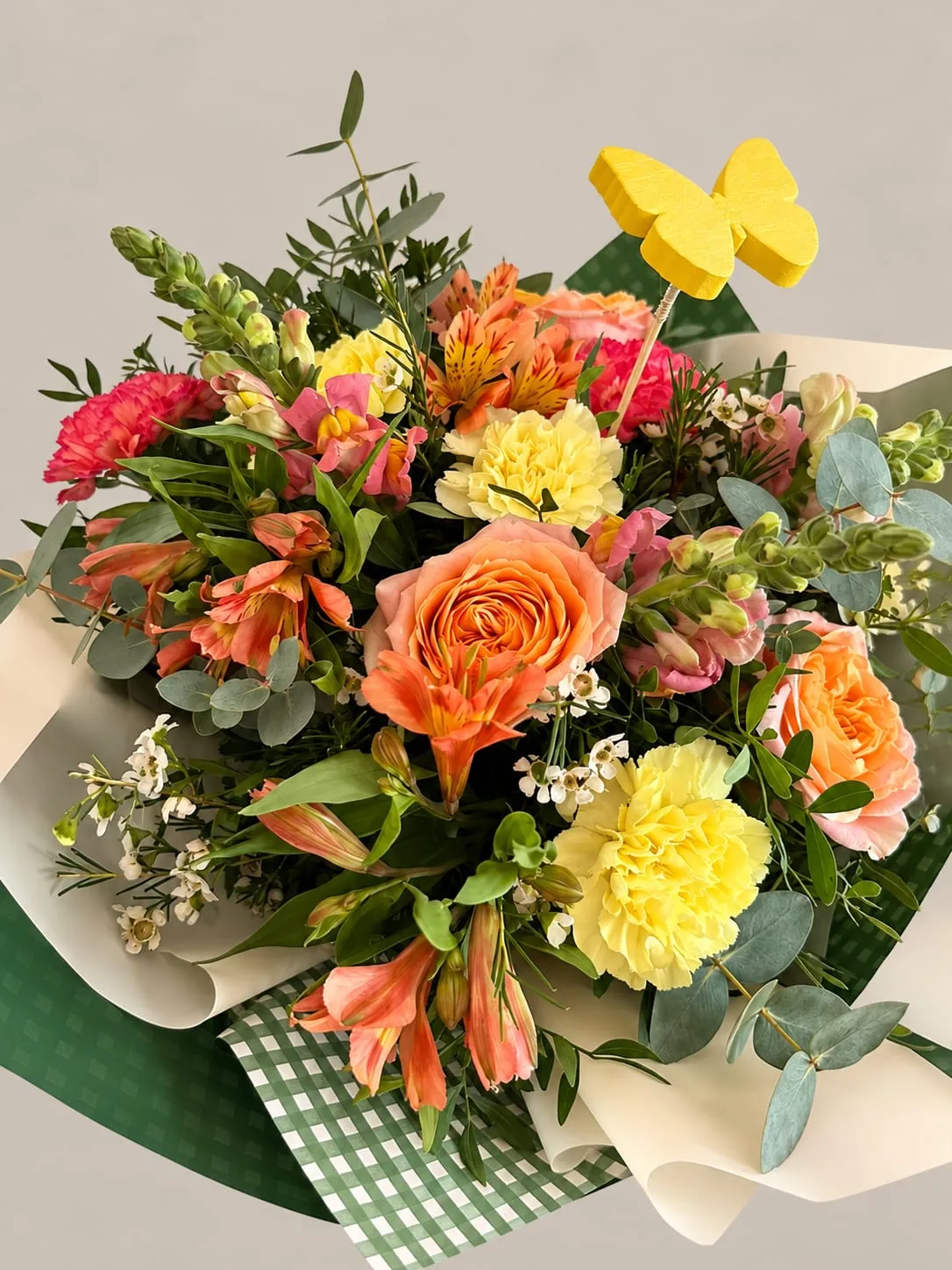 Ramo de rosas, alstroemerias y claveles en tonos naranja, rosa y amarillo con eucalipto, envuelto en papel crema y verde de cuadros, con una mariposa amarilla de adorno.