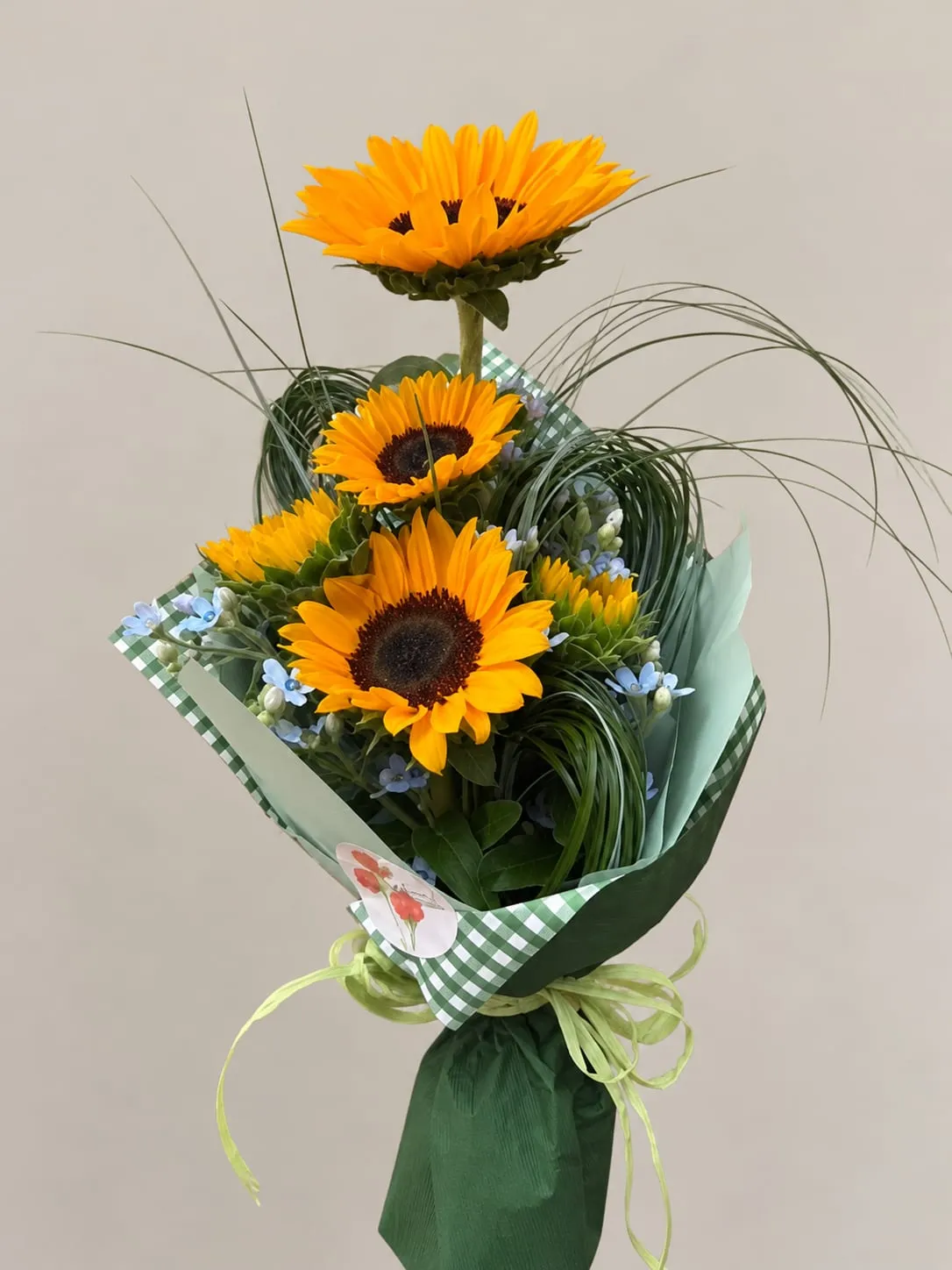 Ramo pequeño de girasoles con flores azules y hojas finas, envuelto en papel verde con lazo de rafia.