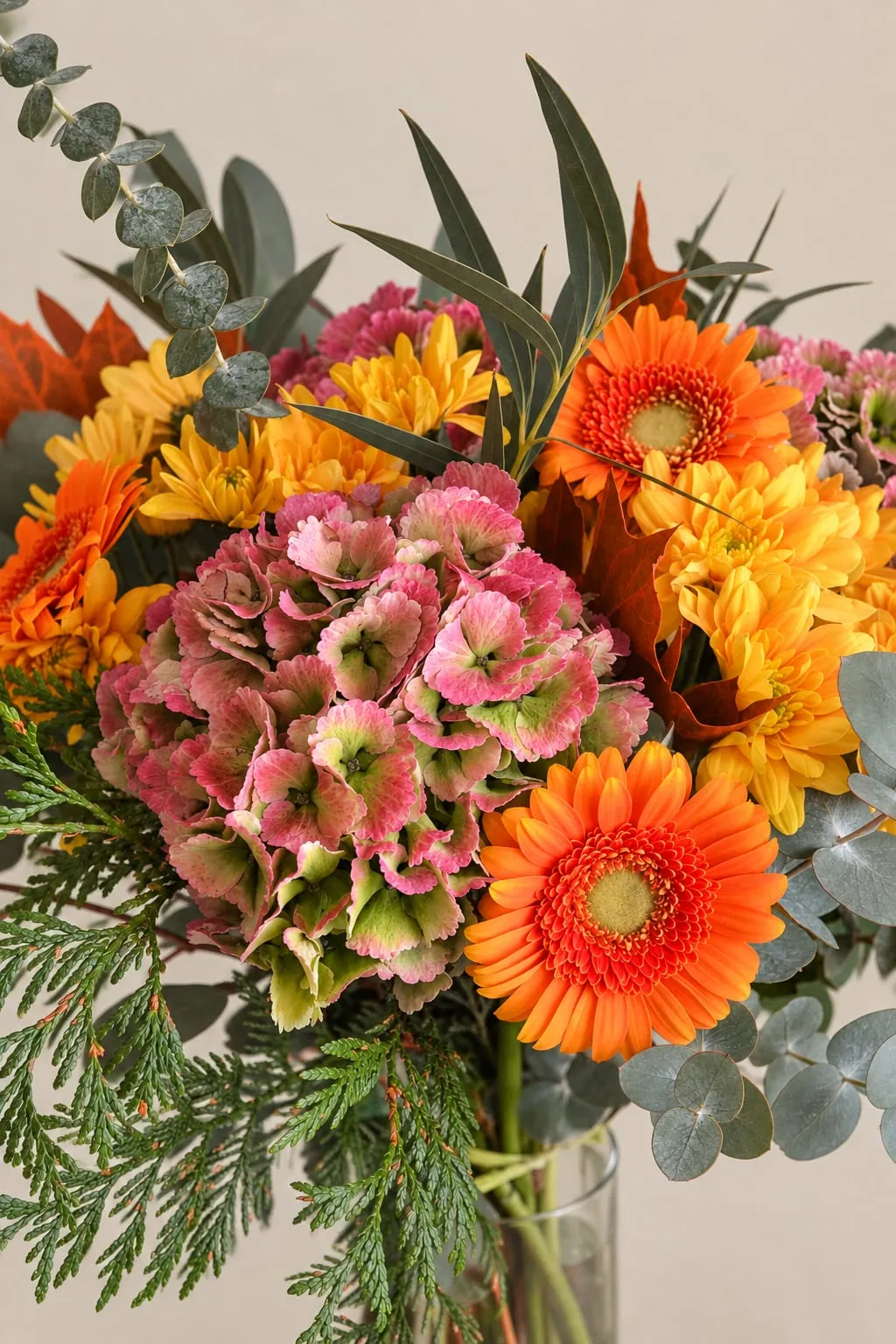 Ramo de hortensias rosadas con gerberas naranjas, crisantemos amarillos, eucalipto y tuya, en un jarrón de cristal.