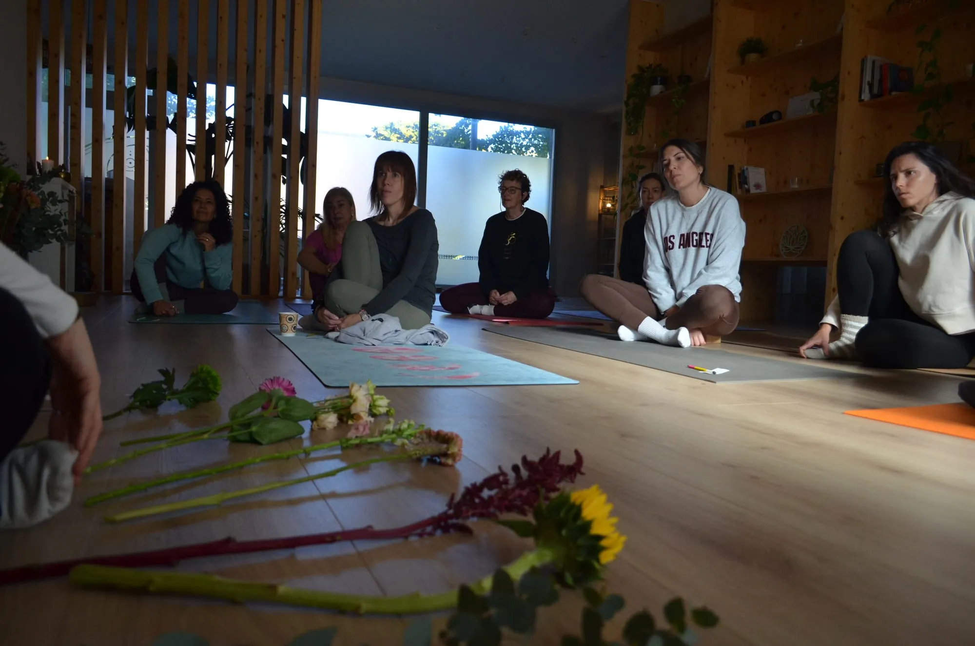 Participantes sentadas en esterillas en una sala de yoga con estanterías de madera, frente a flores sueltas en el suelo: un girasol, amaranto y eucalipto