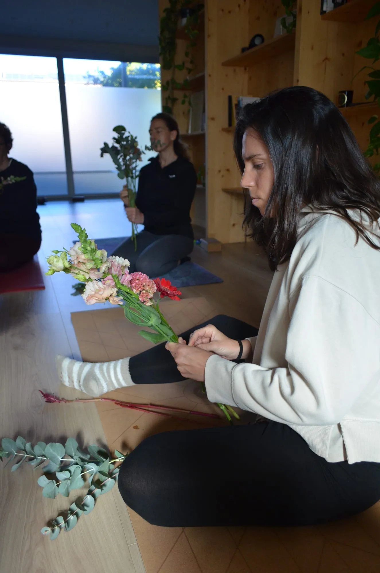 Una participante con jersey blanco monta su ramo de claveles rosas sentada en el suelo, con eucalipto a su lado