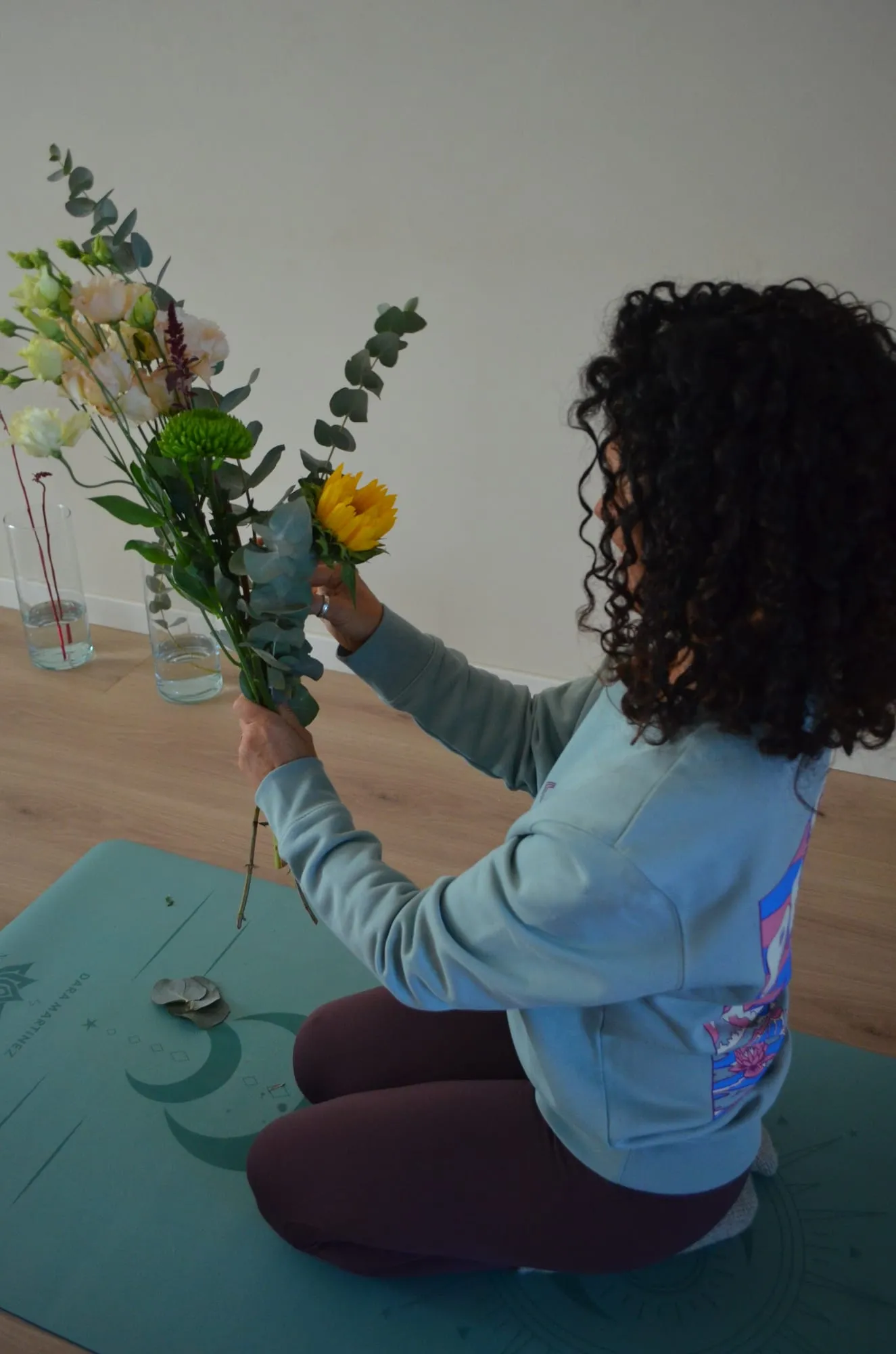 Una participante con sudadera azul, sentada en su esterilla, levanta su ramo con un girasol, alstroemerias y eucalipto
