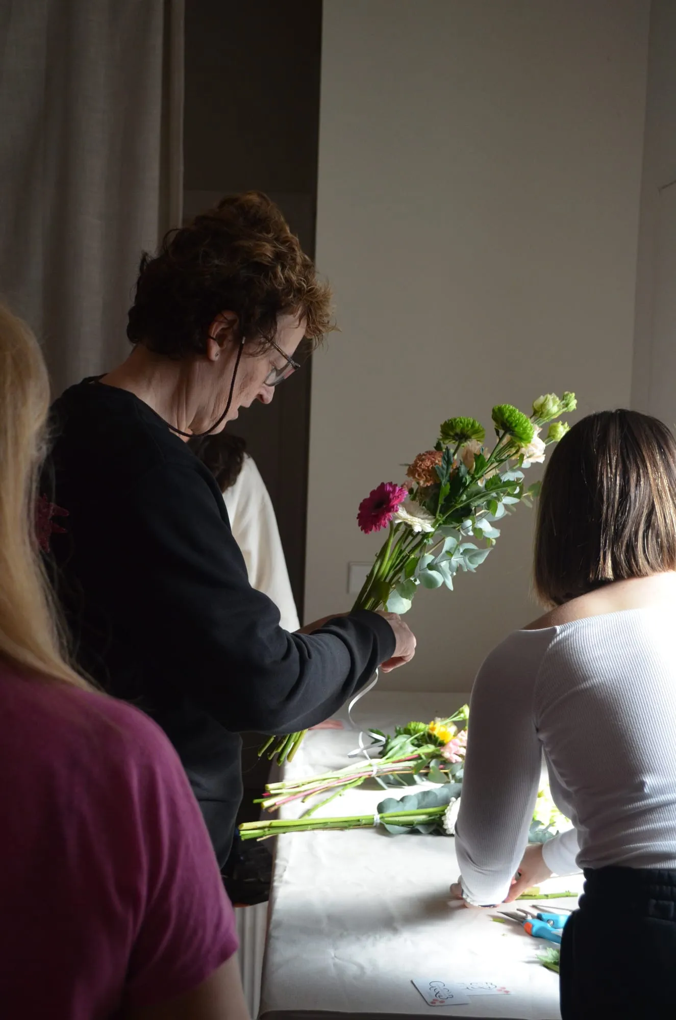 Una participante con gafas sostiene su ramo de gerbera, crisantemos verdes y eucalipto junto a la mesa donde se atan los ramos