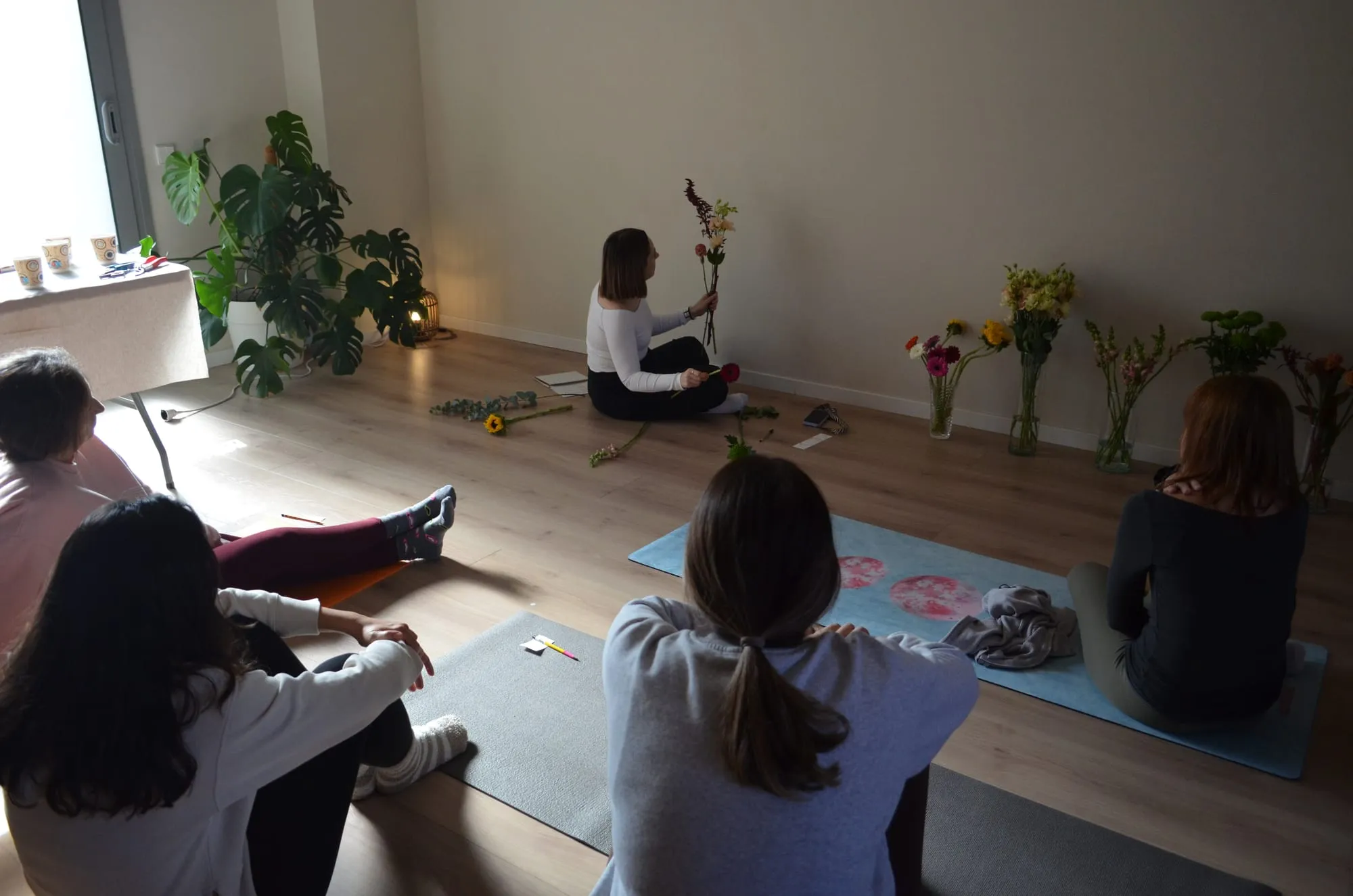 La florista, al fondo de la sala, muestra un tallo a las participantes sentadas en esterillas, con jarrones de flores alineados contra la pared