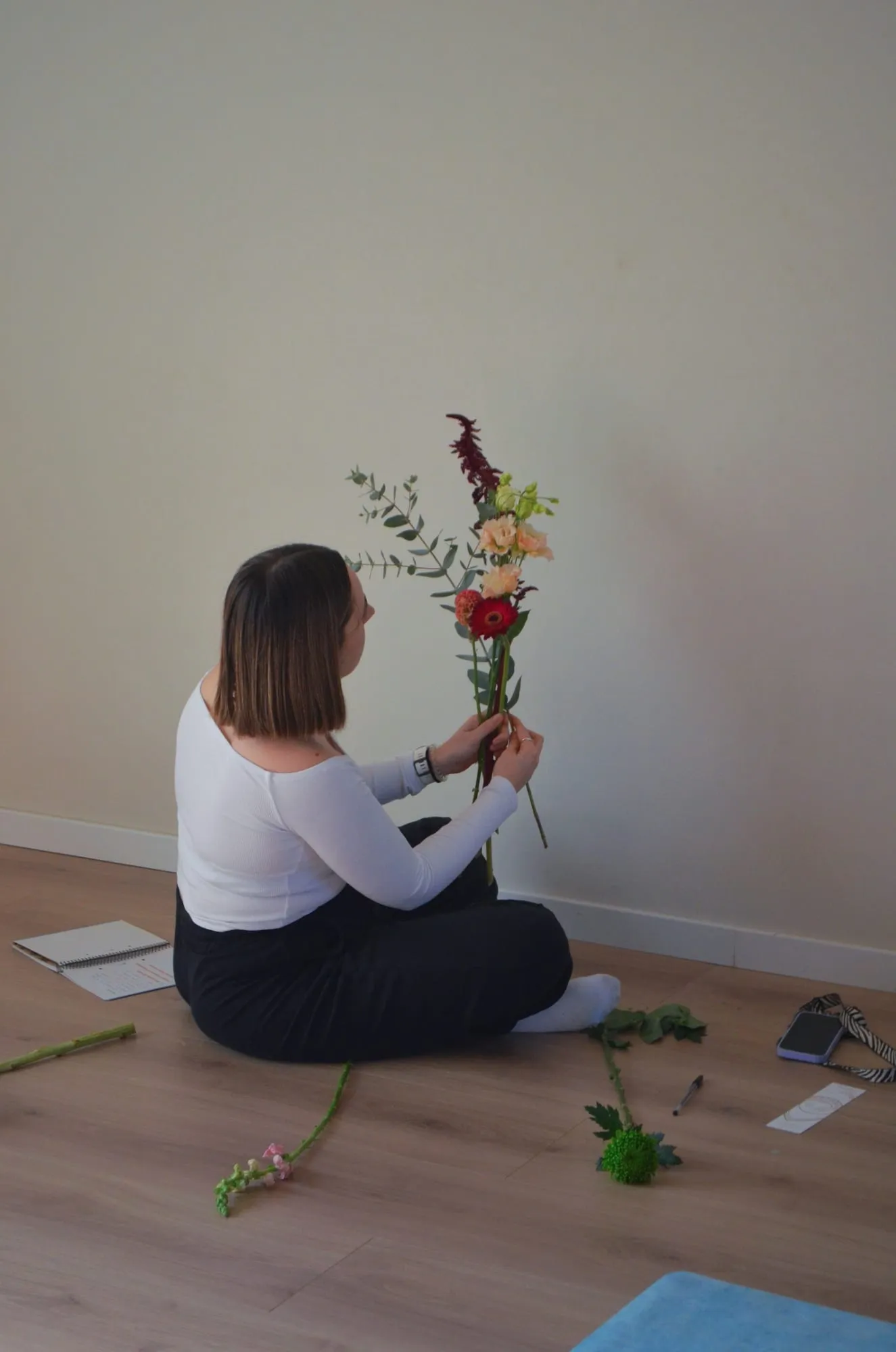 La florista, sentada en el suelo, sostiene un ramo a medio montar con amaranto, rosas y eucalipto, con tallos sueltos a su alrededor