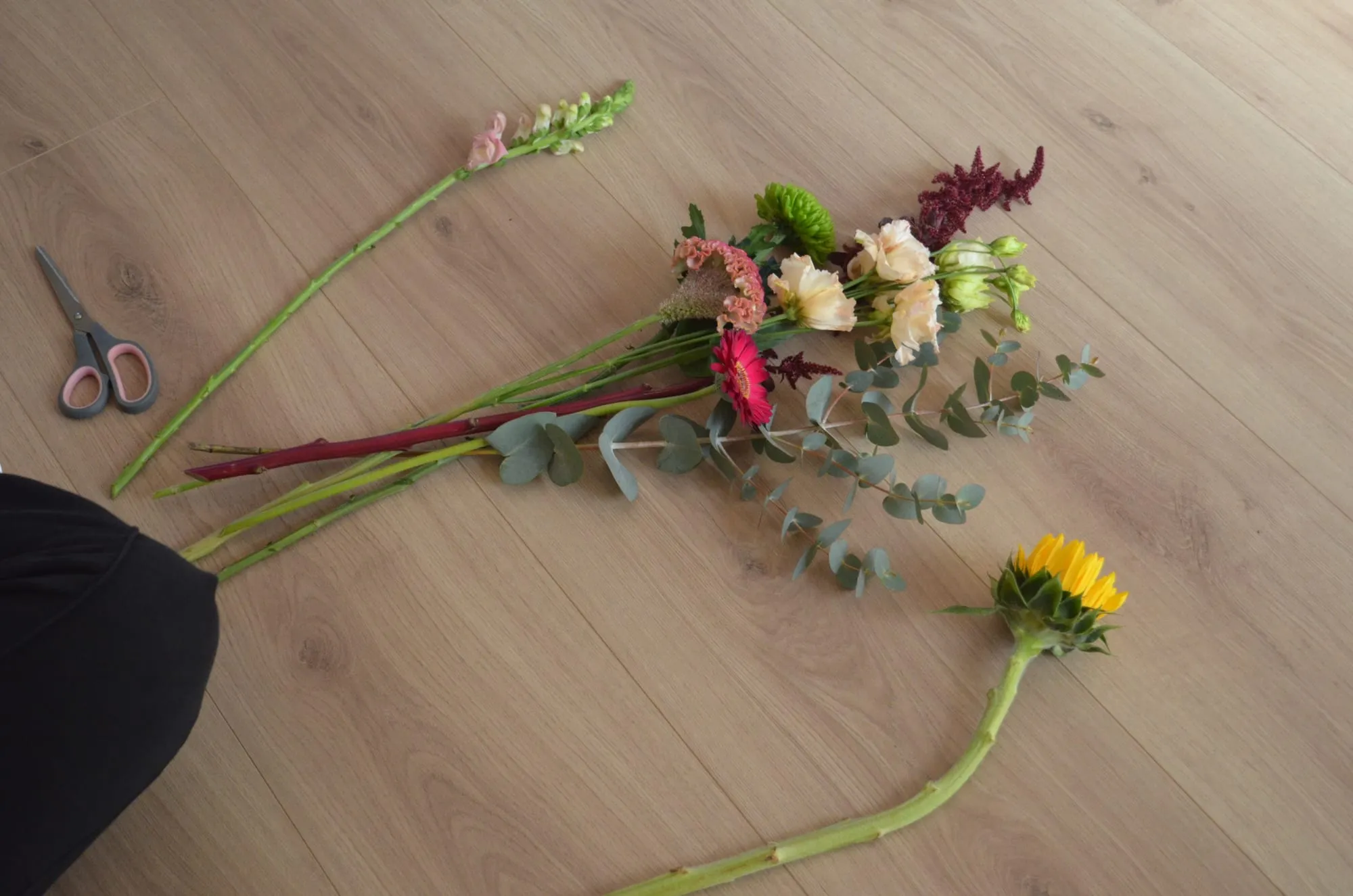 Flores sueltas sobre el suelo de madera: gerbera, amaranto, alstroemerias, boca de dragón, eucalipto y un girasol, junto a unas tijeras