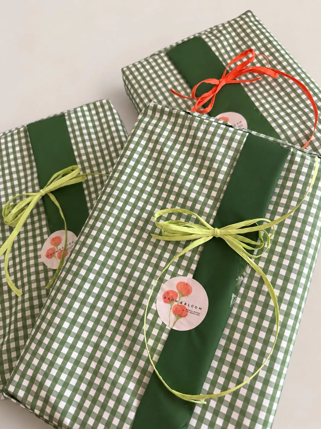 Tres regalos envueltos en papel de cuadros verdes y blancos, con una banda verde, lazos de rafia y una pegatina de flores.